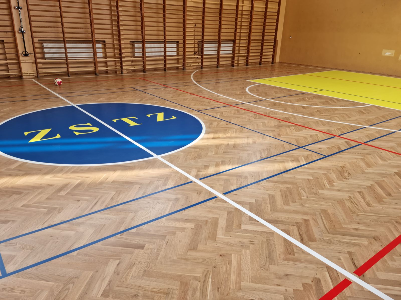 Dębowa jodełka w sali gimnastycznej z logo szkoły i liniami boisk – realizacja Perfekt Parkiet